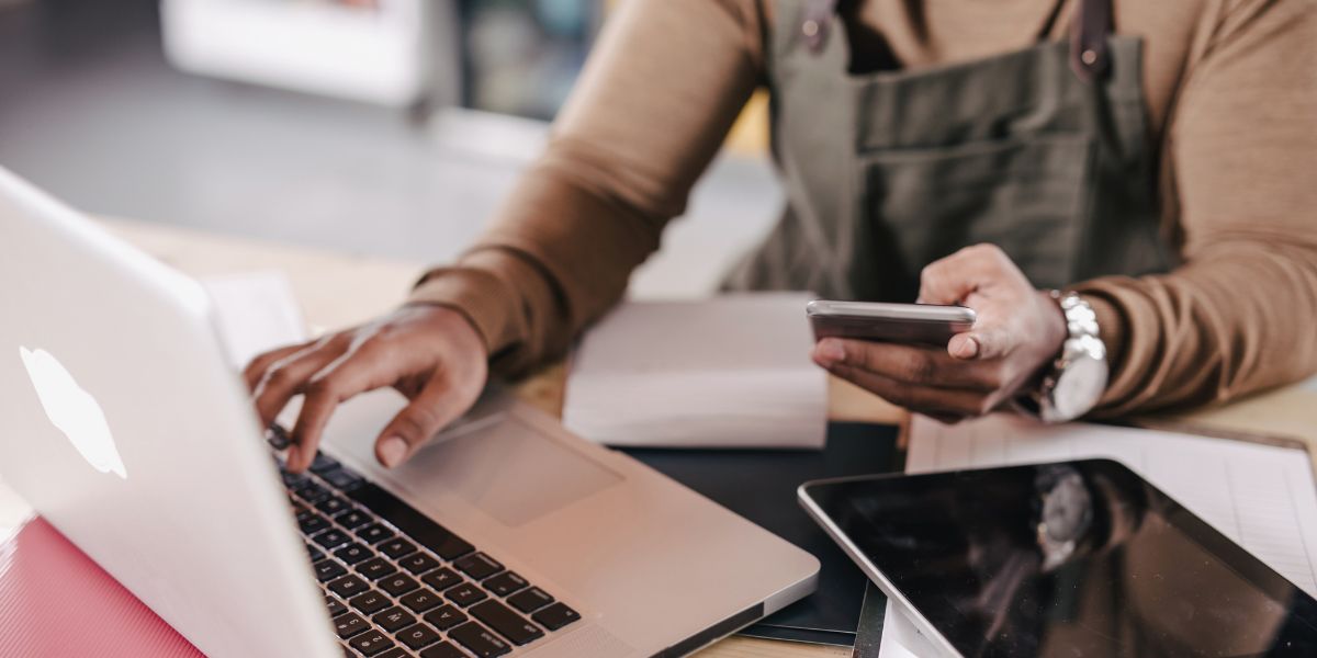 someone in an overall with just body and arms showing looking at a calculator while using a laptop with a tablet also on the desk, used on a blog about the ato small business focus for Q4 2024/25